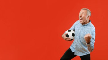 Bir adam elinde futbol topu tutarken gülümsüyor ve kutlama yapıyor. Heyecan ve neşe dolu bir yumruk sallıyor. Arka plan parlak kırmızı ve açık mavi bir gömlek giyiyor..
