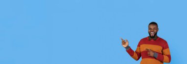 Renkli süveter giyen gülümseyen bir adam boş mavi bir arkaplanı işaret ediyor. Onun neşeli ifadesi heyecan ve pozitifliği iletir. Bu sahne canlı ve çekici bir atmosfer yakalar..