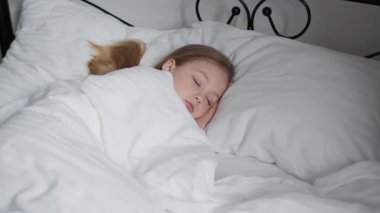 Küçük bir çocuk, yumuşak beyaz battaniyeler içinde, yatağında huzur içinde yatıyor. Güneş ışığı odaya yavaşça süzülür ve sessiz bir sabah boyunca sıcak ve sakinleştirici bir atmosfer yaratır..