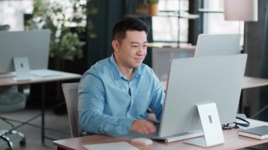 Çağdaş bir ofiste masa başında oturan genç bir adam yazarken bilgisayar ekranına odaklanmış. Güneş ışığı, bitkilerle ve basit dekorlarla dolu çalışma alanını aydınlatır.