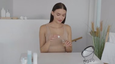 Bir kadın, ışık dolu bir banyodaki basit bir tezgahta oturur ve damlatıcıyı kullanarak derisine serumu dikkatlice uygular. Rahatlatıcı bir atmosferde cilt bakımını seviyor..