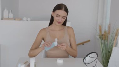 Uzun saçlı bir kadın iyi ışıklandırılmış bir odada bir masada oturuyor. Nazikçe cilt bakım ürünleri uyguluyor..