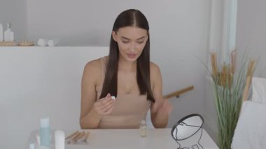 Bir kadın iyi aydınlatılmış bir kibrin önünde oturmuş, dikkatlice cilt bakım ürünlerini uyguluyor. Yer minimalist ve rahatlatıcı, bitki dekoru ve iyi düzenlenmiş güzel eşyalarla dolu..