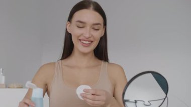 Bir kadın basit bir banyoda elinde pamuk ped ve cilt ürünüyle gülümsüyor. Temiz ürünler ve doğal ışıkla çevrili cilt bakımının keyfini çıkarıyor..