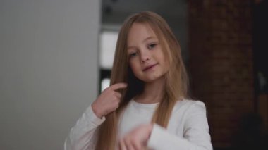 Uzun, düz saçlı genç bir kız şakacı bir şekilde içeride poz veriyor, saçlarına dokunuyor. Yumuşak ışık odayı süzüp sıcak ve davetkar bir atmosfer yaratıyor..