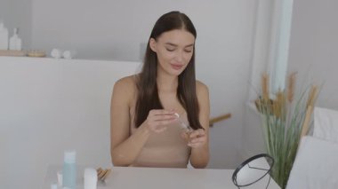 İyi aydınlatılmış bir odada minimalist bir masada oturan bir kadın. Cilt rutinine odaklanmış küçük bir şişeden serum uyguluyor. Doğal ışık sakinleştirici atmosferi güçlendirir..