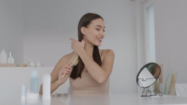 Bir kadın uzun saçlarını temiz, aydınlık bir banyo tezgahında tarıyor. Yuvarlak aynada yansımasına bakarken gülümsüyor. Güzellik ürünleri onun etrafına güzelce yerleştirilmiş..