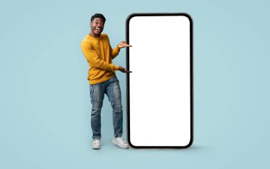 Genç bir Afrikalı Amerikalı adam açık mavi bir arka plana karşı duruyor, büyük bir akıllı telefonu boş bir ekranla sunuyor..