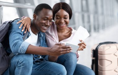 Uçuşu bekliyorum. Havalimanında akıllı telefon kullanan, uçağa binmeden önce terminalde eğlenen, pasaportlarını tutan ve telefon kamerasıyla selfie çeken mutlu Afrikalı Amerikalı çift.