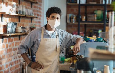 Sosyal uzaklık ve covid-19 salgını sırasında en sevdiği kafe. Milenyum Afrikalı Amerikalı önlüklü ve koruyucu maskeli adam kafeteryanın arkasındaki kahve makinesinin yanında duruyor.
