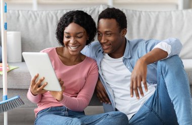 Dijital tabletle gülümseyen aşıklar oturma odasında yerde oturuyor, daire temizleme fikirleri var, kapatıyorlar. Mutlu Afro-Amerikan genç adam ve kadın ev hizmetleri arıyor.