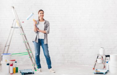 Onarım ve yenileme konsepti. Mutlu genç kadın yeni evi boyuyor, gülümsüyor ve elinde boya ruloları tutuyor.