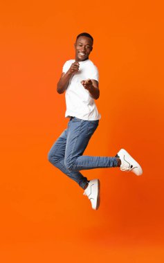 Karizmatik siyahi adam, turuncu stüdyo arka planında zıplarken kamerayı işaret ediyor. Mutlu Afro-Amerikalı genç adam kameraya bakıyor, neşeyle flört ediyor ve zıplıyor.