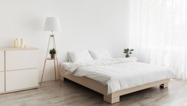 Basit modern tasarım, reklam, teklif. Beyaz yastıklı çift kişilik yatak, yumuşak battaniye, lamba, ahşap zeminde mobilya. Beyaz boş bir duvar, yatak odasının içinde perdeler olan büyük bir pencere.