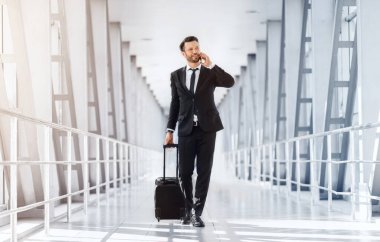 İş gezisi, iş konsepti için seyahat. Bavulu olan yakışıklı girişimci havaalanının önünden geçiyor, cep telefonuyla konuşuyor, pencereden bakıyor, boş alana bakıyor, tam boy çekim yapıyor.