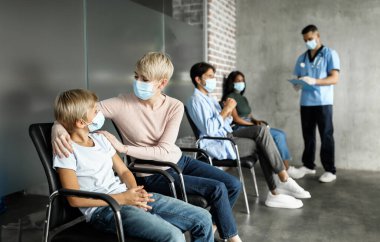 Anne oğlunu teselli ediyor, koruyucu yüz maskeleri takıyor, klinikte gribe karşı aşı oluyor. Çocuklar, yetişkinler aşı olurken salgın konsepti, panorama