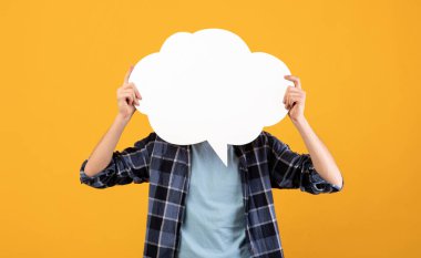 Portakal rengi stüdyo arka planında boş konuşma balonu tutan tanınmamış genç adam tasarım için maket yapıyor. Boş kelime bulutu olan milenyum adamı, reklamın için yer.