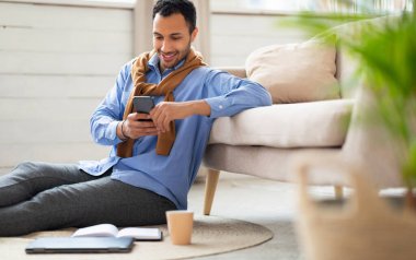 Elinde cep telefonu tutan gülümseyen Orta Doğulu bir adamın portresi oturma odasında koltuğa yaslanmış oturuyor. Tarz sahibi erkek internette geziniyor, internette geziniyor, hücresel uygulama kullanarak video seyrediyor.