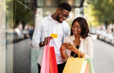 Kolay Ödeme Konsepti. Mutlu Afro-Amerikan erkek ve kadın portresi cep telefonunu birlikte kullanıyor. Kredi kartı ve alışveriş torbaları tutuyor. Alışveriş merkezinin yanında sıradan bir çift bekliyor. Perakende ve Satın Al