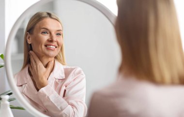 Güzel orta yaşlı kadın aynaya bakıyor ve yumuşak tenine dokunuyor, çekici olgun bayan yansımaya gülümsüyor, evde yaşlanma karşıtı cilt rutininin tadını çıkarıyor, seçici odaklanıyor.