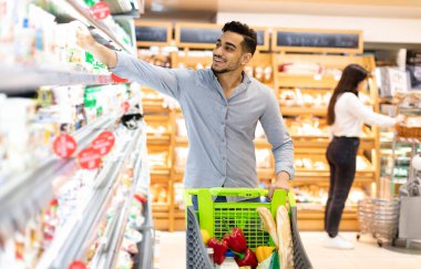 Market alışverişi. Modern süpermarkette raftan mandıra ürünü alan Arap adam. Müşteri seçiyor ve alışveriş yapıyor. Dükkanda bakkalla dolu market arabasıyla ayakta duruyor.