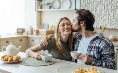 Yüzlerce yıldır gülümseyen beyaz koca, sakallı sarılıp karısını yanağından öpüyor ve modern mutfak içinde kahve içiyor. Boş zaman, içkili kahvaltı ve evde günaydın.