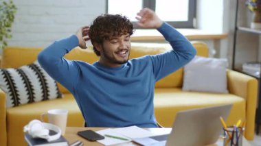 Bir erkek, parlak bir oturma odasında bir masada otururken gülümsüyor ve kollarını uzatıyor. Sıradan mavi bir kazak giyerek bir video görüşmesiyle meşgul..