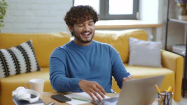 Kıvırcık saçlı genç bir adam parlak bir oturma odasında oturur ve dizüstü bilgisayarıyla meşgul olur. Neşeli ve odaklanmış görünüyor. Kanepesi ve sanat eserleri olan rahat bir atmosferle çevrili..