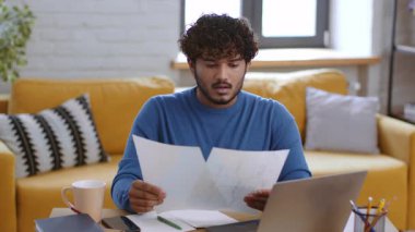 Bir erkek, rahat bir oturma odasındaki masada dokümanları inceliyor. Doğal ışık pencerelerden süzülür, dizüstü bilgisayarda çalışırken ve elinde kağıtlar tutarken konsantrasyonunu vurgular..