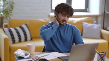 Kıvırcık saçlı genç bir erkek parlak bir oturma odasında bir masada oturuyor. Boynunu ovalarken dizüstü bilgisayarına odaklanmış gibi görünüyor. Yakınlarda bir fincan kahve ve defter var..