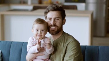 Neşeli bir baba gülümseyen bebeğini parlak ve modern bir oturma odasında tutar. Babanın sakalı var ve bebek alkışlıyor, sıcak ve sevgi dolu bir atmosfer yaratıyor..