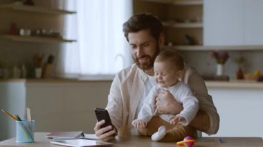 Bir baba parlak bir mutfakta masasında otururken bebeğiyle mutlu bir şekilde iletişim kurar. Bebek gülümsüyor ve babasının telefonuna uzanıyor. Atmosfer sıcak ve neşeli..