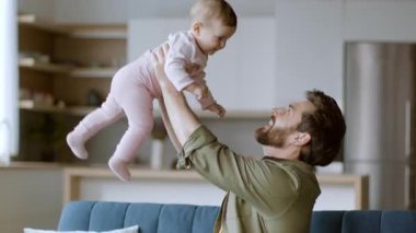 Bir baba bebeğini neşeli bir şekilde doğal ışıkla dolu, modern bir oturma odasında kaldırır. Neşeli an, sevgi dolu bir aile ortamı sergileyerek bağlarını ve mutluluklarını yakalar..