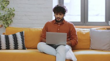 Kıvırcık saçlı genç bir adam sarı bir kanepede otururken dizüstü bilgisayarına odaklanır. Odanın büyük pencere ve bitkileri var, sıcak ve davetkar bir atmosfer yaratıyor..