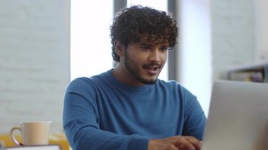 Kıvırcık saçlı genç bir adam iyi ışıklandırılmış bir odada bir masada oturur. O, yakınlarda bir fincan kahve içerken dizüstü bilgisayarına odaklanmış. Atmosfer rahat ve verimli..