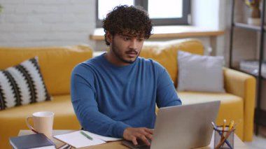 Kıvırcık saçlı bir adam rahat bir oturma odasında dizüstü bilgisayarında yazı yazıyor. Doğal ışık pencereden girer, sıcak bir atmosferle uzayı aydınlatır..