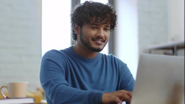 Genç bir adam dizüstü bilgisayarı açık bir masada oturuyor, daktilo ediyor ve gülümsüyor. Doğal ışıkla dolu parlak ve davetkar bir odada kitaplarla çevrili sıcak bir içecekten hoşlanır..