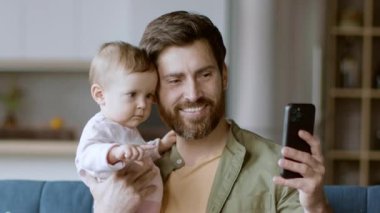Bir baba bebeğini telefonuna bakarken kucağında tutar. Doğal ışıkla dolu sıcak bir oturma odasında video görüşmeleri sırasında aynı gülüşü paylaşıyorlar. Atmosfer sıcak ve neşeli..