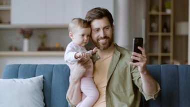 Bir baba video görüşmesi sırasında bebeğini tutarken gülümser. İyi ışıklandırılmış bir salonda rahat bir koltukta oturuyorlar ve birlikte kaliteli vakit geçiriyorlar..