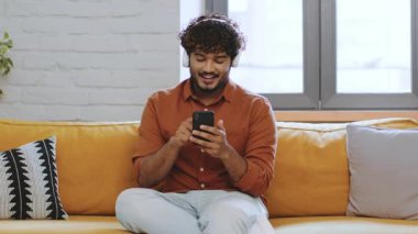 Kıvırcık saçlı genç bir adam canlı bir kanepede rahat bir şekilde oturur ve akıllı telefonuna gülümseyerek bakar. Güneş ışığı pencereden içeri akar ve odada sıcak bir atmosfer yaratır..