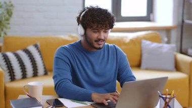 Genç bir adam ahşap bir masada oturuyor, dizüstü bilgisayarına odaklanmış. Kulaklık takıyor ve yakınlarda kahve içiyor..