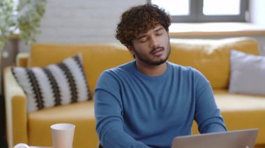 Bir erkek, parlak bir oturma odasında dizüstü bilgisayarına odaklanarak rahatça oturur. Güneş ışığı pencereden süzülüp sıcak bir atmosfer yaratır. Rahat bir sarı kanepe ve saksı bitkisi rahatlatıcı bir hava katar..