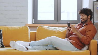 Bir erkek, parlak sarı bir kanepede rahat bir şekilde oturuyor, akıllı telefonuyla meşgul. Rahat ve memnun görünüyor, şık bir oturma odasında, pencerelerden yansıyan doğal ışıkla..