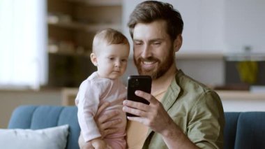 Bir baba bebeğini kollarında tutarken ikisi de akıllı telefona birlikte bakar. Yumuşak mobilyalarla çevrili, doğal ışıkla dolu, rahat bir oturma odasında oturuyorlar..