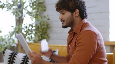 Genç bir adam aydınlık bir odada rahatça oturup tabletle etkileşime geçiyor. Bitkiler ve rahat mobilyalarla çevrili bir şekilde elinde içki tutarken odaklanmış ve rahat görünüyor..