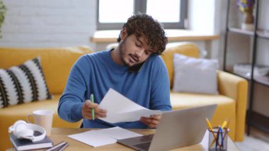 Kıvırcık saçlı bir adam ev ofisinin ahşap masasında oturur. Önemli evrakları incelerken telefonla konuşuyor. Yakınlarda bir fincan kahve duruyor, odaklanmış bir çalışma ortamı yaratıyor..