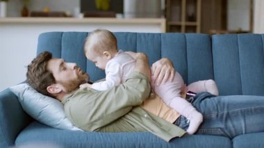 Bir baba bebeğiyle oynarken rahat bir kanepede yatar. Konforlu oturma odasında yumuşak ışıklandırma ve modern dekor var. Bu aile zamanlarında kahkaha ve neşeyi paylaşıyorlar..