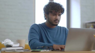 Genç bir adam parlak ve modern çalışma alanında dizüstü bilgisayarına odaklanıyor. Kitaplar, kulaklıklar ve kahve fincanlarıyla dolu bir masada oturuyor..