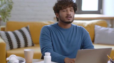 Bir erkek parlak bir oturma odasında otururken dizüstü bilgisayarına konsantre oluyor. Mavi bir kazak giyiyor ve derin düşünce belirtileri gösteriyor. Mekan yakınlarda rahat bir kanepe ile aydınlık..