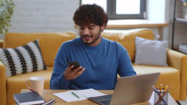 Bir erkek parlak bir oturma odasında ahşap bir masada oturuyor, bir dizüstü bilgisayara odaklanırken akıllı telefon kullanıyor. Doğal ışık boşluğu doldurarak üretkenlik için rahat bir ortam yaratıyor..